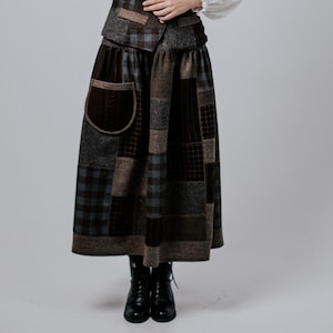 May include: A woman wearing a white blouse with a brown and blue patchwork vest and skirt. The skirt has a large patch pocket on the front.