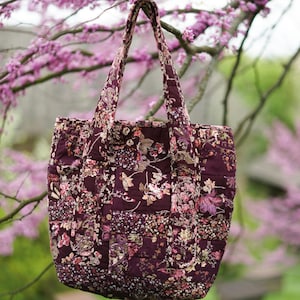 Peut inclure: Un sac fourre-tout matelassé avec un motif floral dans les tons bordeaux, rose et crème. Le sac a deux anses et est suspendu à une branche d'arbre. Le design du sac présente un style patchwork avec une variété d'imprimés floraux.