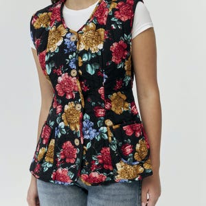 Könnte beinhalten: Schwarze Steppweste mit einem lebendigen Blumenmuster in Rot, Gelb und Blau. Die Weste hat einen V-Ausschnitt, Holzknöpfe und zwei Vordertaschen. Darunter wird ein weißes T-Shirt getragen. Ein stilvolles Oberbekleidungsstück.