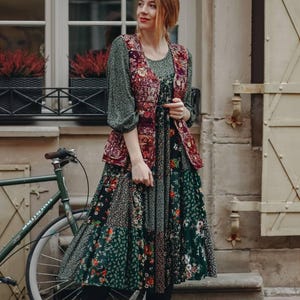 Può includere: Una donna indossa un vestito in patchwork verde e rosso con stampa floreale e un gilet bordeaux. È in piedi accanto a una bicicletta e guarda di lato.