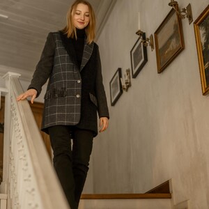 Puede incluir: Una persona desciende una escalera de madera, vistiendo una blazer a cuadros gris oscuro y negro con detalles de flecos. También lleva un cuello alto negro y pantalones negros. El fondo presenta obras de arte enmarcadas y apliques de pared.