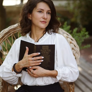 May include: A woman in a white blouse and black skirt sits in a wicker chair holding a closed black book with gold lettering that reads "ЗОЛОТЕ ГЛОГО" (Golden Hawthorn).