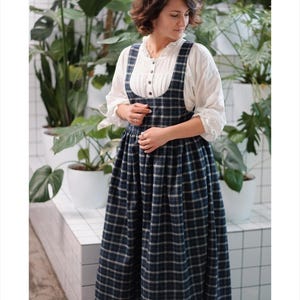 May include: A woman wearing a white blouse with a ruffled collar and a navy blue plaid jumper dress. The dress has a full skirt and suspender straps. The background features potted plants and a white tiled wall.