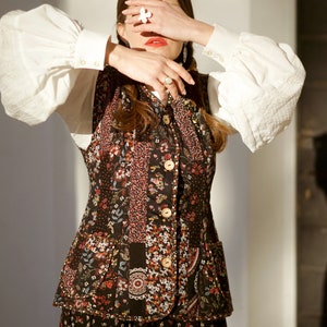 May include: A woman wearing a white blouse with puffed sleeves and a black floral vest over a black floral skirt. She is covering her eyes with her hand and wearing a ring on her finger.