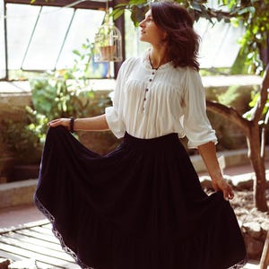 May include: A woman wearing a white blouse with a button-up front and a black tiered skirt with lace trim. She is standing in a greenhouse with plants and a birdcage in the background.