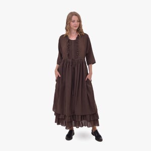 Pode incluir: Um vestido marrom de manga comprida com decote com babados e saia em camadas. O vestido apresenta cintura com cord&atilde;o, bolsos e &eacute; combinado com sapatos pretos. O vestido &eacute; feito de um tecido macio e fluido.