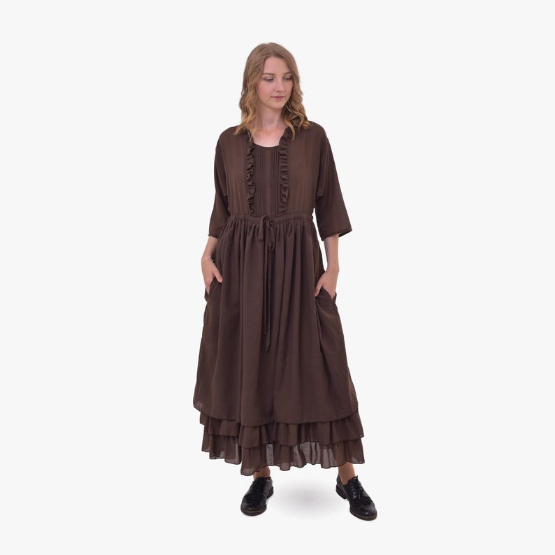 Pode incluir: Um vestido marrom de manga comprida com decote com babados e saia em camadas. O vestido apresenta cintura com cord&atilde;o, bolsos e &eacute; combinado com sapatos pretos. O vestido &eacute; feito de um tecido macio e fluido.
