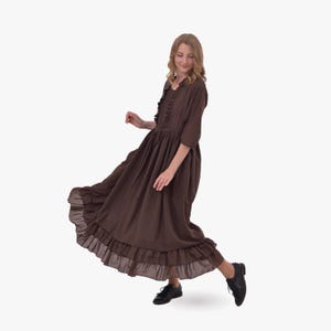 Pode incluir: Um vestido longo e esvoa&ccedil;ante castanho com acabamento de babados e mangas tr&ecirc;s quartos. O vestido tem cintura alta e decote com babados. A pessoa est&aacute; usando sapatos pretos. O vestido parece ser feito de um tecido leve.