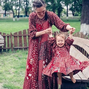 May include: A woman and a child in matching red floral dresses. The woman is holding the child's hands, lifting them up. The dresses have long sleeves and a patchwork design. The background features a wooden fence, trees, and green grass.