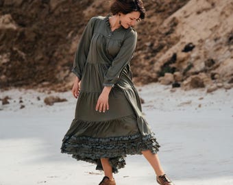 May include: An olive green maxi dress with long sleeves and a ruffled hem. The dress features a high neckline and is paired with brown leather boots. The background shows a sandy, natural environment.