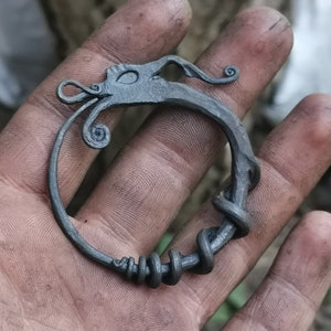 May include: A hand holding a metal dragon-shaped ring with a coiled tail. The ring is made of dark metal and has a detailed design.