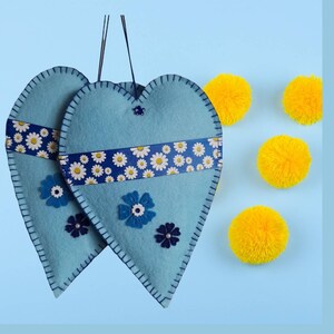 May include: Two pale blue felt heart decorations with daisy ribbon and blue flower details. The hearts are stitched with dark blue thread and have black hanging strings. There are yellow pom-poms scattered around.
