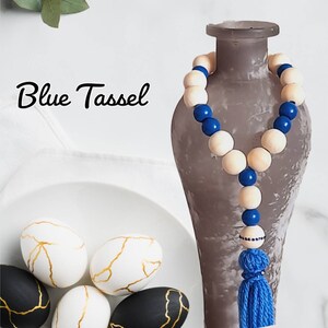May include: A decorative beaded garland with alternating natural wood and blue beads, topped with a blue tassel. The text "Blue Tassel" is written in cursive. Decorative eggs with gold crackle designs are in the foreground.