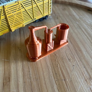 May include: A copper-colored model of a moonshine still with a large copper pot, a smaller copper barrel, and a copper pipe connecting the two. The still is sitting on a wooden surface.