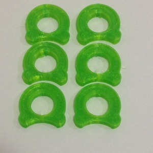 May include: Six translucent green, ring-shaped objects with a decorative bottom design are arranged in two rows of three. The objects have a layered appearance and a circular opening in the centre. The background is plain white.