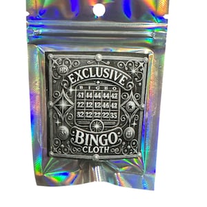 May include: A silver and black bingo card with the words "Exclusive Bingo Cloth" printed on it. The card is in a small, iridescent, zippered bag.