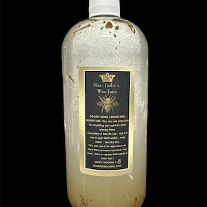 May include: A clear glass bottle with a white cap filled with a light brown liquid. The bottle has a gold label with the text "Mrs. Judie's Woo Juice" and instructions for use. The label also features a bee and a crown.