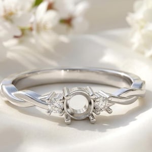 May include: A silver engagement ring with a twisted band design. The ring features a central setting for a gemstone, flanked by two smaller, round-cut accent stones. The ring is displayed on a white surface with blurred white flowers in the background.