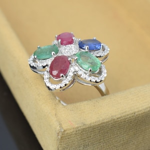 Puede incluir: Anillo de plata con diseño floral, engastado con cinco piedras preciosas ovales: dos rubíes, dos esmeraldas y un zafiro. Cada gema está rodeada por un halo de pequeños diamantes brillantes. El anillo se muestra sobre un fondo neutro.