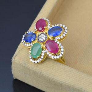 May include: A gold-toned ring with a flower design. The ring features oval-cut gemstones in red, blue, and green, surrounded by small white stones. The center of the flower has a cluster of small white stones.