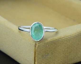 May include: A silver ring featuring a faceted, oval-cut emerald gemstone. The emerald has a light green hue and is set in a simple, polished silver band. The ring is displayed on a neutral-toned surface.