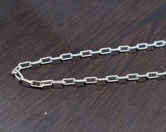 May include: A silver chain necklace with a box link design. The chain is lying flat on a dark brown surface.