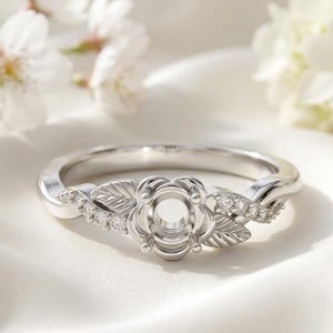 May include: A silver engagement ring with a floral design. The ring features a flower-shaped setting and leaf accents, with small diamonds set along the band. The ring is displayed on a white surface with soft lighting.