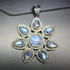 May include: A silver pendant with a starburst design featuring seven moonstone gems. The pendant is set with small clear stones around each moonstone.
