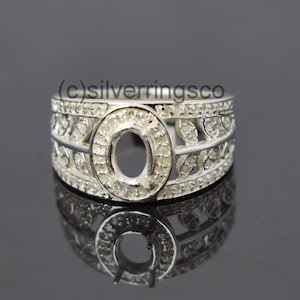 May include: A silver ring with a round, open setting for a gemstone. The ring is decorated with a leaf design and small, clear stones.