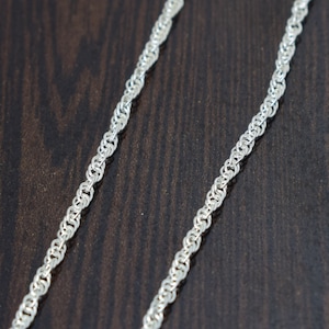 May include: A silver chain necklace with a delicate, interwoven design. The chain is made of small, linked circles that create a smooth, flowing look.