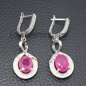 May include: A pair of silver dangle earrings with a pink oval gemstone surrounded by smaller clear gemstones. The earrings have a simple, elegant design.