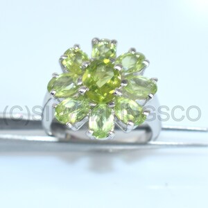 May include: A silver ring with a floral design, set with light green peridot gemstones. The oval-cut stones are arranged in a cluster, creating a flower-like pattern. The ring's band is polished, and the gemstones are set closely together, creating a vibrant design.