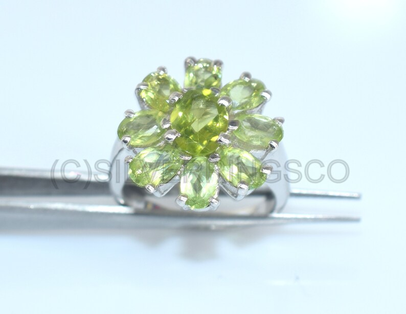 May include: A silver ring with a floral design, set with light green peridot gemstones. The oval-cut stones are arranged in a cluster, creating a flower-like pattern. The ring's band is polished, and the gemstones are set closely together, creating a vibrant design.