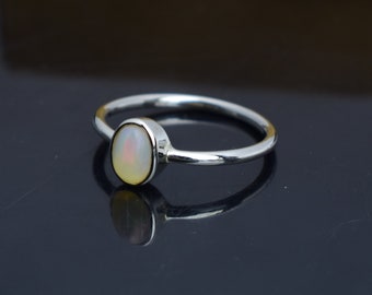 Pode incluir: Um anel de prata com uma pedra de opala oval. A opala tem uma base branca cremosa com flashes de amarelo e rosa. A banda do anel é fina e polida, refletindo a luz.