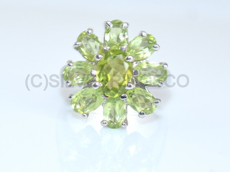 May include: A silver ring with a floral design, set with oval-cut peridot gemstones. The ring's setting and band are silver. The peridot stones are a bright, light green, arranged in a flower shape with a central, larger stone. The ring is a piece of jewellery.