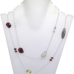 May include: A silver necklace with two strands, featuring a variety of gemstones. The necklace includes large, irregularly shaped red stones, smaller brown and yellow stones, and a few blue and iridescent stones. Displayed on a white bust.