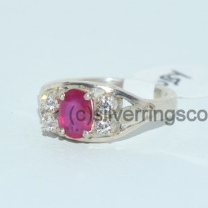 May include: A silver ring featuring a red oval gemstone surrounded by smaller white gemstones. The ring is displayed against a white backdrop.