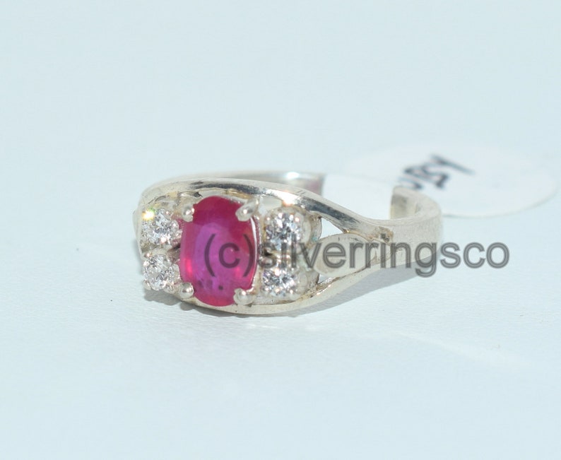May include: A silver ring featuring a red oval gemstone surrounded by smaller white gemstones. The ring is displayed against a white backdrop.