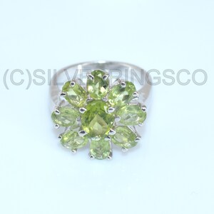 May include: A silver ring with a floral design, set with oval-cut, light green peridot gemstones. The ring's band is polished, and the gemstones are arranged around a central stone. The background is bright.