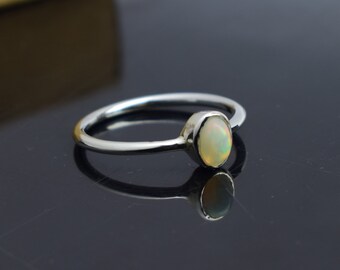 Pode incluir: Um anel de prata com uma pedra preciosa de opala oval. A opala tem uma cor de base branca leitosa com flashes de cores iridescentes. A banda do anel é fina e polida, refletindo a luz.