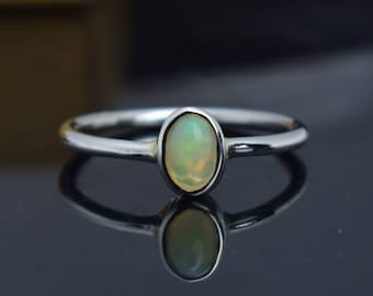 Pode incluir: Um anel de prata com uma pedra preciosa de opala oval. A opala apresenta uma cor pálida e iridescente com tons de verde e amarelo. A banda do anel é fina e polida, refletindo a luz. A pedra preciosa é engastada numa moldura, realçando a sua forma e cor.