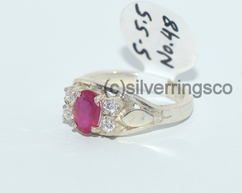 May include: A silver ring featuring a red oval gemstone surrounded by smaller white gemstones. The ring is marked with the number "848" and the letters "S.S.S."