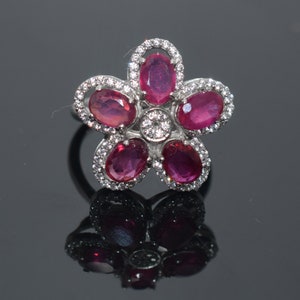 以下が含まれることがあります： 5つの赤い宝石が、より小さな白い宝石で囲まれた花のデザインが施された銀の指輪です。