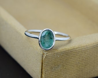 May include: A silver ring featuring an oval-cut, emerald-green gemstone. The gemstone is set in a bezel setting, and the band is thin and polished. The ring is displayed on a neutral-toned surface, highlighting the vibrant green of the stone.