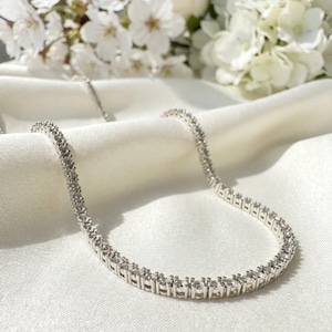 Puede incluir: Un collar de tenis en tono plateado que presenta una hilera continua de piedras redondas, transparentes y facetadas engastadas en un marco metálico. El collar está dispuesto en forma curva, mostrando el tamaño uniforme y el brillo de cada engaste.