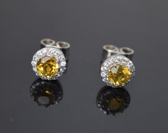 May include: A pair of silver stud earrings with a round yellow gemstone surrounded by a halo of smaller white gemstones.