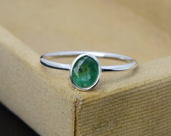 May include: A silver ring featuring a vibrant green emerald gemstone. The emerald is oval-shaped and set in a simple, elegant band. The ring is displayed on a neutral-toned surface, highlighting the gemstone's color and clarity.