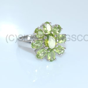 May include: A silver ring with a floral design, set with oval-cut, light green gemstones. The gemstones are arranged in a petal-like pattern around a central stone. The ring has a polished finish and is displayed against a light background.