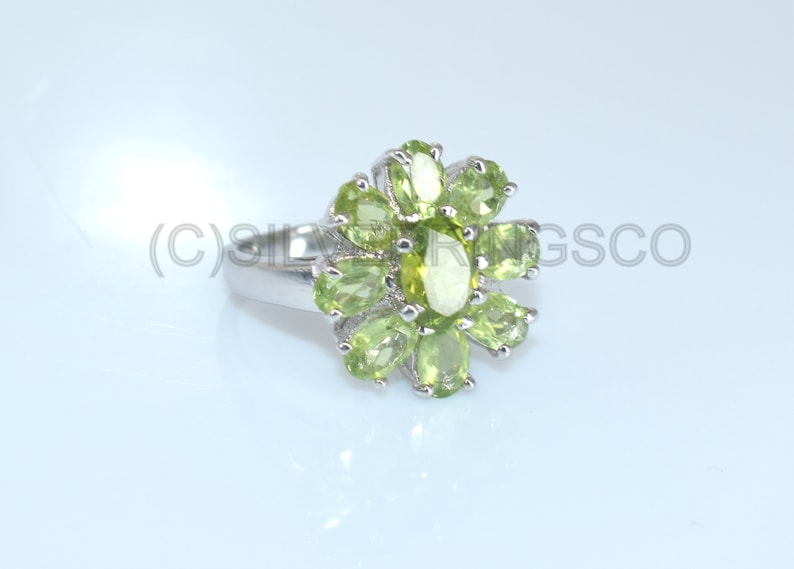 May include: A silver ring with a floral design, set with oval-cut, light green gemstones. The gemstones are arranged in a petal-like pattern around a central stone. The ring has a polished finish and is displayed against a light background.