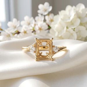 May include: A gold ring featuring a rectangular prong setting designed to hold a center stone. The band is accented with a single small round brilliant-cut diamond on each side of the central mount, showcasing a polished metallic finish.
